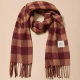A plaid scarf in shades of burgundy and tan, draped in a loop on a solid beige background. A small white square label with the brand name 'theca' is visible on the right side of the scarf.