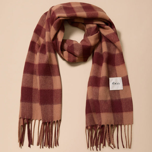 A plaid scarf in shades of burgundy and tan, draped in a loop on a solid beige background. A small white square label with the brand name 'theca' is visible on the right side of the scarf.