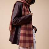 A woman wearing a brown and burgundy plaid scarf with fringe, styled over a dark brown faux-leather jacket and white pants, set against a solid beige studio background.
