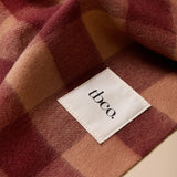 A close-up of a soft plaid blanket or scarf in muted burgundy, dusty rose, and tan tones, with a white fabric label reading 'tbco.' sewn onto one section.