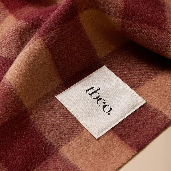 A close-up of a soft plaid blanket or scarf in muted burgundy, dusty rose, and tan tones, with a white fabric label reading 'tbco.' sewn onto one section.