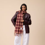 A model stands against a solid beige studio background wearing a dark brown leather jacket, white wide-leg trousers, black shoes, and a rust-brown and beige plaid scarf.
