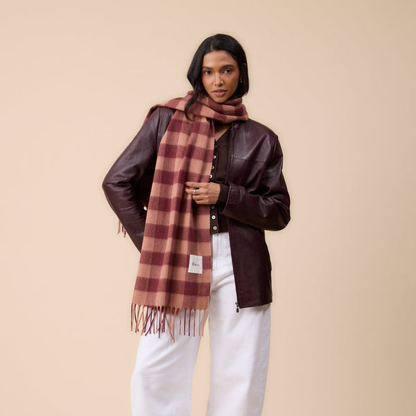 A model stands against a solid beige studio background wearing a dark brown leather jacket, white wide-leg trousers, black shoes, and a rust-brown and beige plaid scarf.