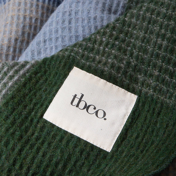 A close-up of a textured waffle-knit blanket in dark green and light blue, with a white fabric label stitched onto the green section that reads 'tbco.'.