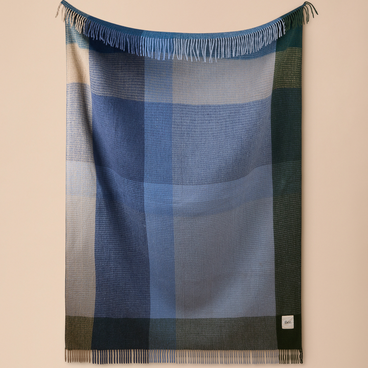 A plaid fringed throw blanket in shades of blue, green, and beige, hanging on a neutral beige wall.