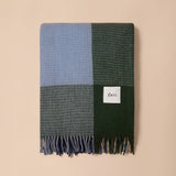 A folded knit throw blanket with a four-block pattern in light blue, dark green, and gray, finished with fringed edges and a small white label reading 'thea'.