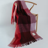 A woven throw blanket with a red, burgundy, and dusty pink plaid pattern and fringed edges, draped over a light wood chair against a plain light gray background.