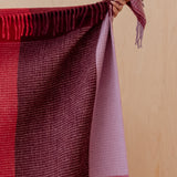 A close-up of a woven throw blanket with fringe, held by a hand against a light wood panel background. The blanket features a color-blocked pattern of deep burgundy, dark red, and dusty lavender-purple.
