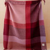 A plaid throw blanket with fringed edges, featuring a color-blocked pattern in shades of pink, red, and purple, displayed against a light wood background.