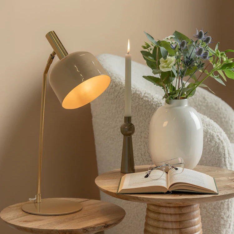 Lamp, candle, vase with flowers, and open book on a wooden table against a beige wall.