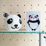 A white paper lantern shaped like a panda's head sits on a glass shelf next to a printed card featuring a black-and-white panda with pink cheeks, set against a light wood pegboard background.