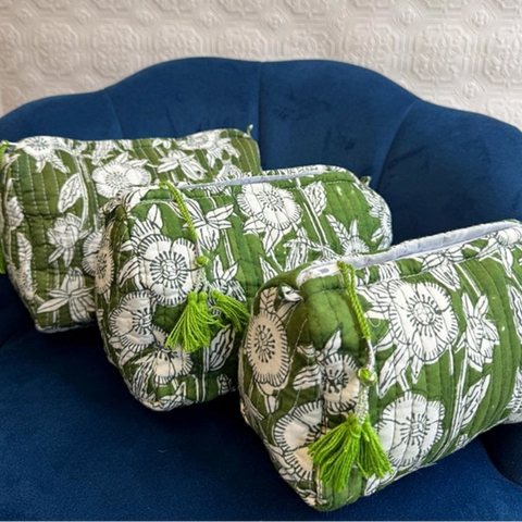 Three quilted cosmetic pouches with a green and white floral print, green tassels, and silver zippers, arranged on a dark blue velvet armchair.