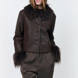 A model wearing a dark brown faux-leather jacket with a large dark brown fur collar and matching dark brown trousers, photographed against a light gray studio background.