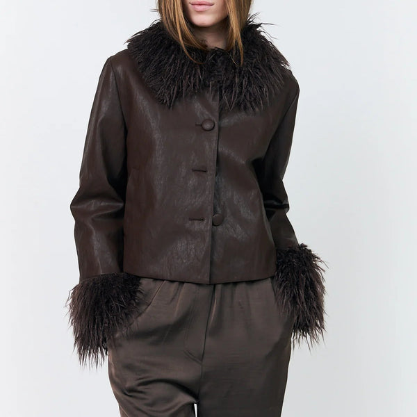 A model wearing a dark brown faux-leather jacket with a large dark brown fur collar and matching dark brown trousers, photographed against a light gray studio background.