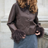A woman wearing a brown faux-leather jacket with dark brown fur trim at the collar and cuffs, paired with gray trousers, standing against a stone wall.