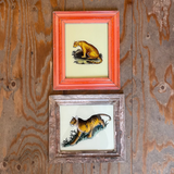 Two vintage animal paintings in frames hanging on a rustic wood-paneled wall. The top painting features a yellow and black cat in a bright red-orange frame, and the bottom painting shows a tiger in a distressed brown frame.