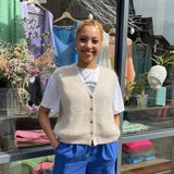 Person wearing a beige cardigan, white shirt, blue shorts, and red shoes standing in front of a store with clothing displayed.