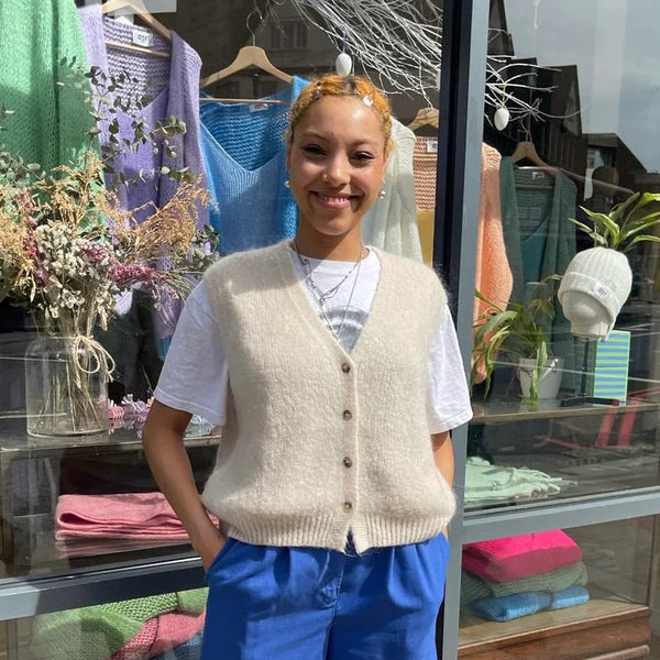 Person wearing a beige cardigan, white shirt, blue shorts, and red shoes standing in front of a store with clothing displayed.