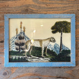 A framed vintage needlepoint artwork depicting a cheetah in a landscape, mounted on a rustic wooden wall.