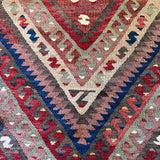A close-up of a traditional geometric kilim rug featuring nested diamond and chevron patterns in red, blue, beige, gray, and brown.