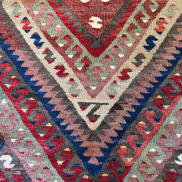 A close-up of a traditional geometric kilim rug featuring nested diamond and chevron patterns in red, blue, beige, gray, and brown.