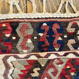 Close-up of a woven rug with a geometric tribal pattern in red, navy blue, beige, and brown, featuring a thick braided tassel fringe at the top.
