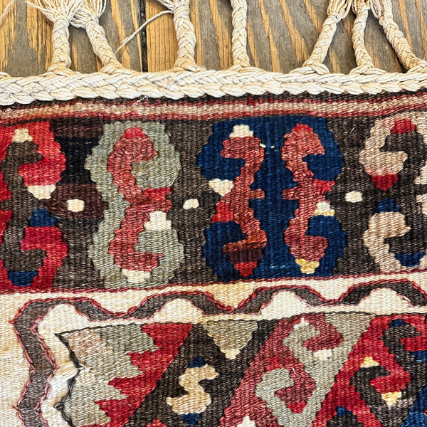 Close-up of a woven rug with a geometric tribal pattern in red, navy blue, beige, and brown, featuring a thick braided tassel fringe at the top.
