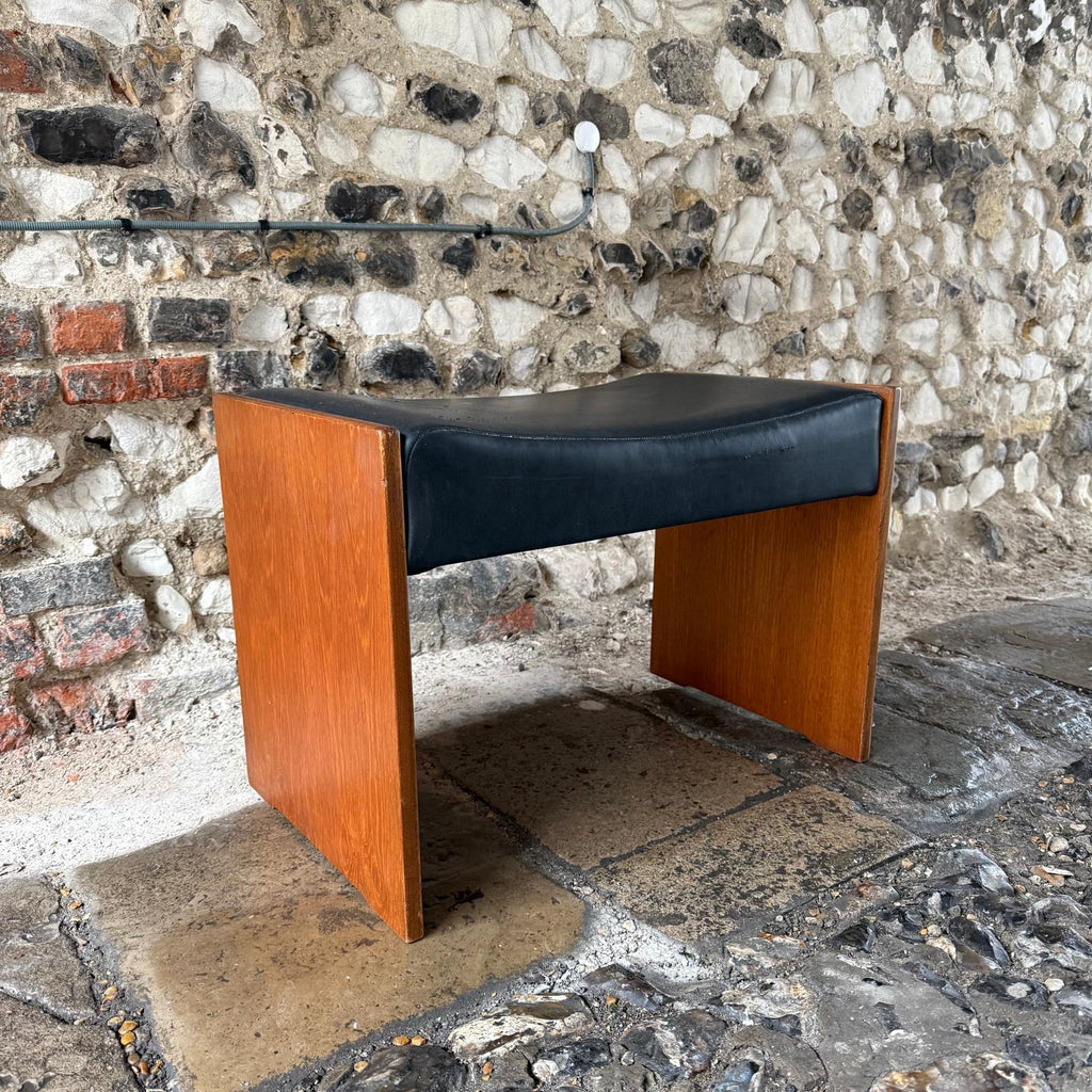 Wooden stool with black cushion against a stone wall.