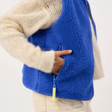 A close-up of a person wearing a bright royal blue sherpa fleece vest over a cream knit sweater and light pants, with a hand reaching into a side zipper pocket.