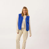 A woman modeling a bright blue textured fleece vest over a cream knit sweater and cream pants, standing against a plain white studio background.