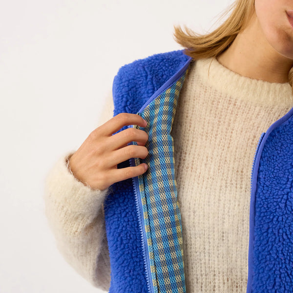 Close-up of a person wearing a bright blue fleece vest over a cream knit sweater, holding the vest open to show a patterned blue and yellow lining.