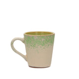 A handmade ceramic mug with a textured, speckled finish in cream and light green, shown against a plain white background.