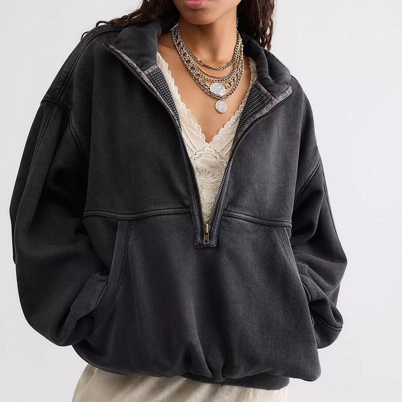 A woman wearing a dark charcoal gray half-zip pullover hoodie with a half-placket, layered over a cream lace top and skirt, against a light gray studio background.