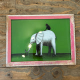 A framed wall art piece featuring a stylized white elephant with a black trunk and a small black bird on its back, set against a bright green background. The frame is distressed pink and white, displayed on a rustic wooden plank wall.