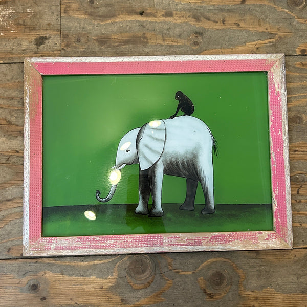 A framed wall art piece featuring a stylized white elephant with a black trunk and a small black bird on its back, set against a bright green background. The frame is distressed pink and white, displayed on a rustic wooden plank wall.