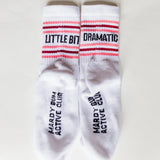 A pair of white crew socks laid flat on a light background. The socks feature pink and dark red stripes near the cuff with the words 'LITTLE BIT DRAMATIC' in black, and the words 'MARDY BUN ACTIVE CLUB' printed vertically on the soles.
