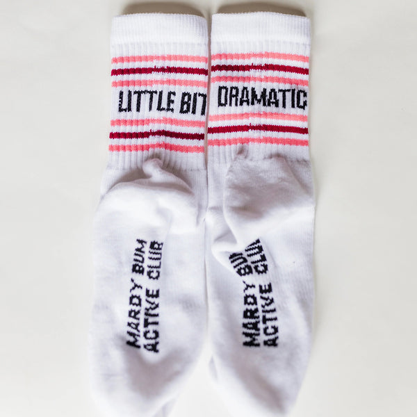 A pair of white crew socks laid flat on a light background. The socks feature pink and dark red stripes near the cuff with the words 'LITTLE BIT DRAMATIC' in black, and the words 'MARDY BUN ACTIVE CLUB' printed vertically on the soles.
