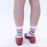 White crew socks with pink and dark red stripes and black text reading 'BITTLE BASTARD' and 'DRAMATIC', worn with red ballet flats on a white background.