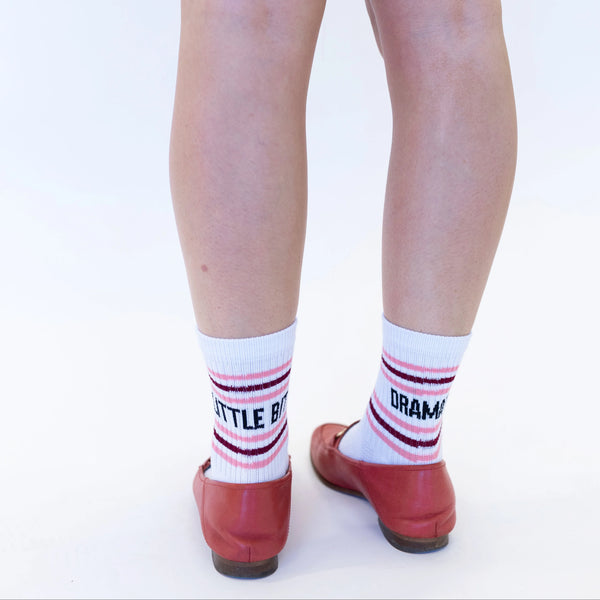 White crew socks with pink and dark red stripes and black text reading 'BITTLE BASTARD' and 'DRAMATIC', worn with red ballet flats on a white background.