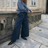 A woman stands outdoors on a stone terrace in front of a classical building, wearing a blue and white vertically striped long-sleeve top, navy wide-leg trousers, and black pointed shoes.