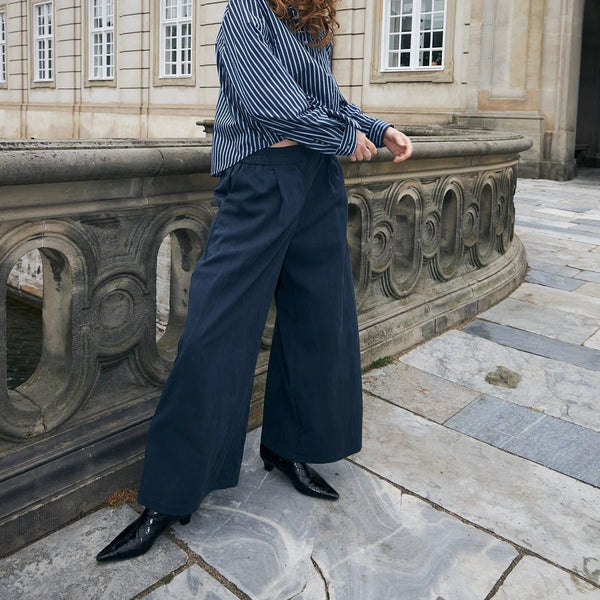 A woman stands outdoors on a stone terrace in front of a classical building, wearing a blue and white vertically striped long-sleeve top, navy wide-leg trousers, and black pointed shoes.