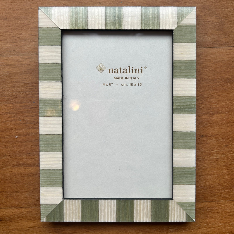 A rectangular picture frame with a striped border in sage green, cream, and white, displayed on a brown wooden surface. The center insert shows the brand name 'natalini'.