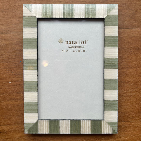 A rectangular picture frame with a striped border in sage green, cream, and white, displayed on a brown wooden surface. The center insert shows the brand name 'natalini'.