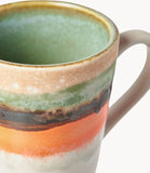 A close-up of a ceramic mug with a handle, featuring horizontal bands of mint green, beige, dark brown, orange, and white glaze on a white background.