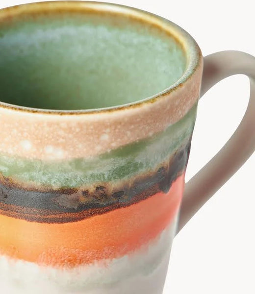 A close-up of a ceramic mug with a handle, featuring horizontal bands of mint green, beige, dark brown, orange, and white glaze on a white background.