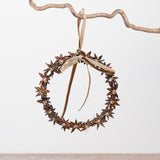 A decorative hanging wreath made from brown star anise flowers with a beige ribbon bow, suspended from a rustic branch against a plain light background.