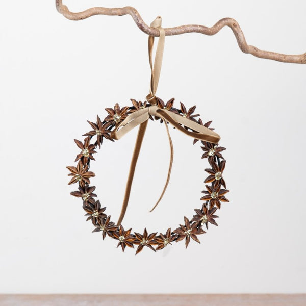 A decorative hanging wreath made from brown star anise flowers with a beige ribbon bow, suspended from a rustic branch against a plain light background.