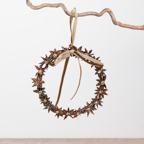 A decorative hanging wreath made from brown star anise flowers with a beige ribbon bow, suspended from a rustic branch against a plain light background.