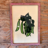 A framed art print featuring green seed pods and leaves on a cream background, displayed on a rustic wooden table. The photo is taken from above and is captured on a desktop screen with a macOS interface visible.